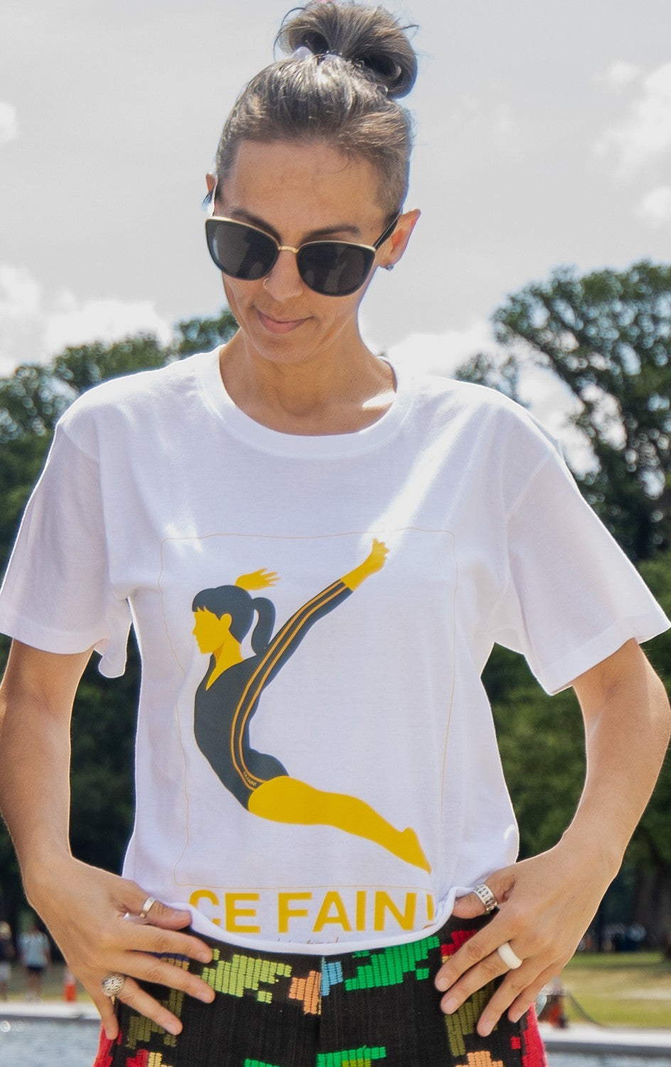 Ce Fain! Nadia Tshirt in white cotton featuring a yellow and black gymnast graphic, worn by a woman with sunglasses.